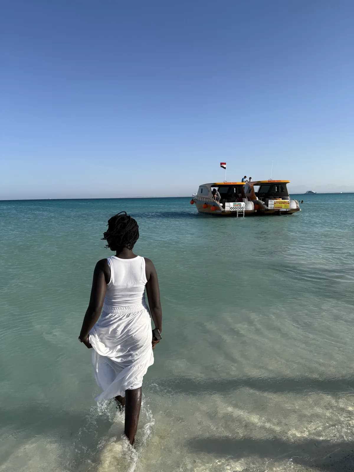 Frau im weißen Kleid geht durch türkisfarbenes Wasser auf zwei gelbe Wassertaxis zu, an einer Insel vor Hurghada